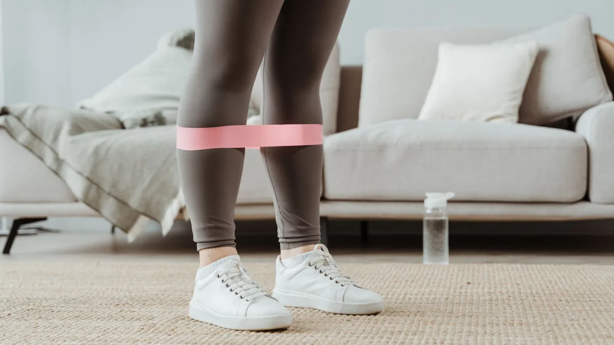 Jambes d'une personne avec une bande élastique rose autour des cuisses, devant un canapé