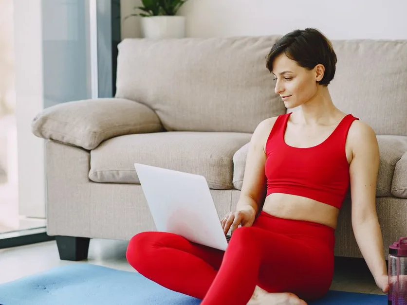 Femme en tenue de sport assise sur un tapis près du canapé, qui consulte un ordinateur portable après sa séance