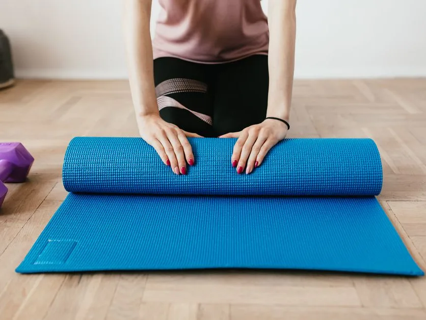 Femme qui déroule un tapis de sport bleu au sol, à côté de deux haltères, dans un salon baigné de lumière