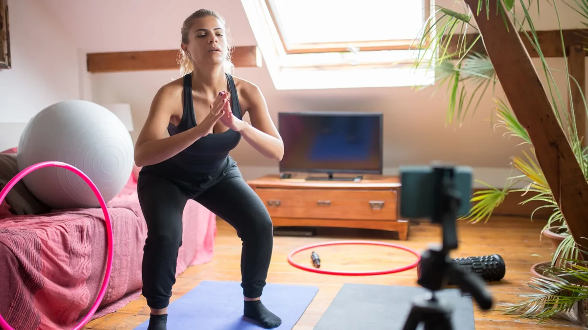 Femme qui fait un squat sur un tapis dans une chambre, un téléphone sur trépied la filme