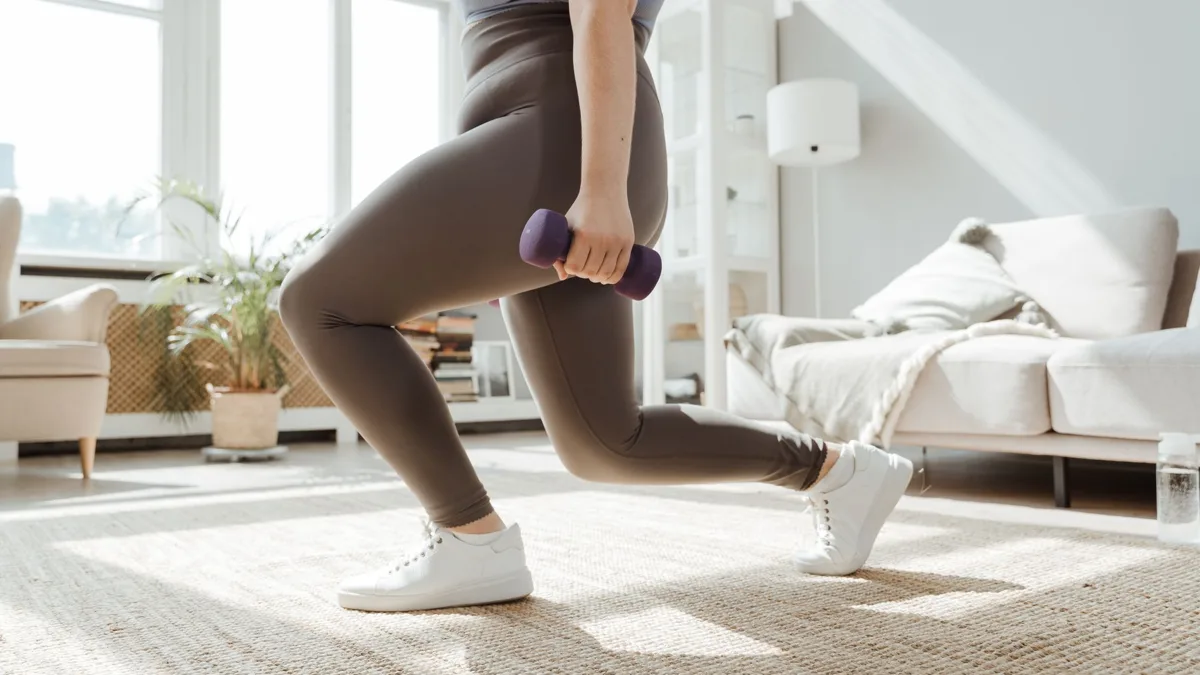 Jambes d'une femme en fente avant, un haltère violet à la main, dans un salon clair