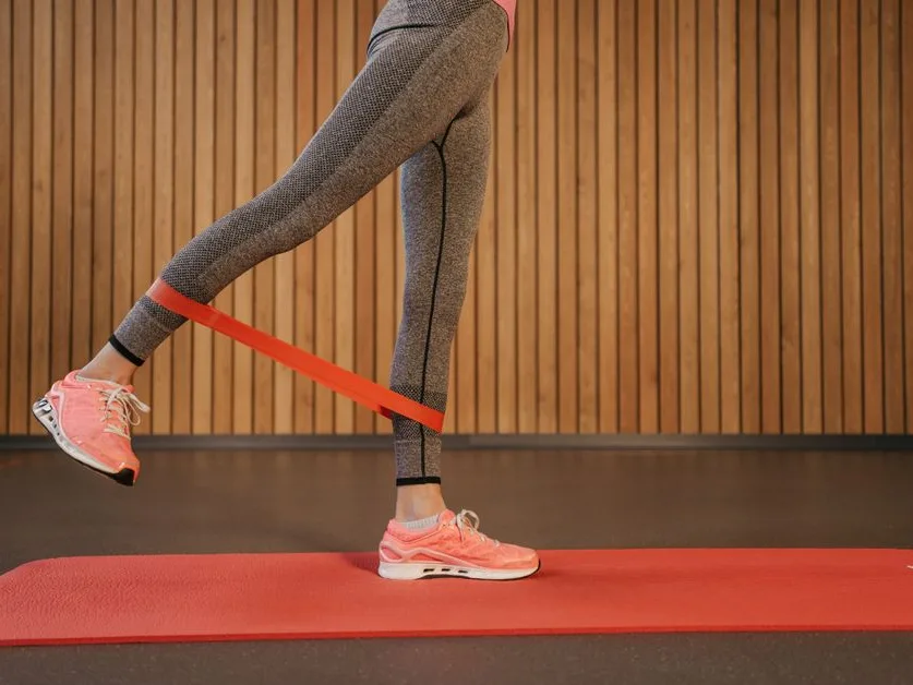 Femme qui travaille les jambes avec un élastique rouge sur un tapis de sol, dans une pièce de la maison